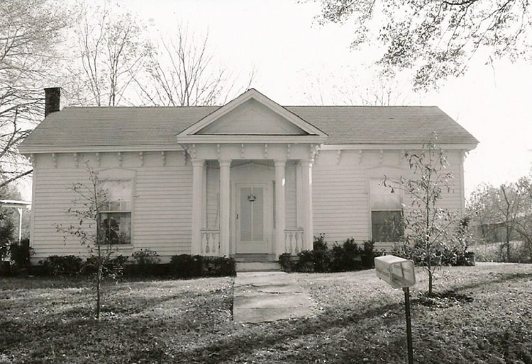 Front view of single-story house with four front columns sidewalk and mailbox in the foreground