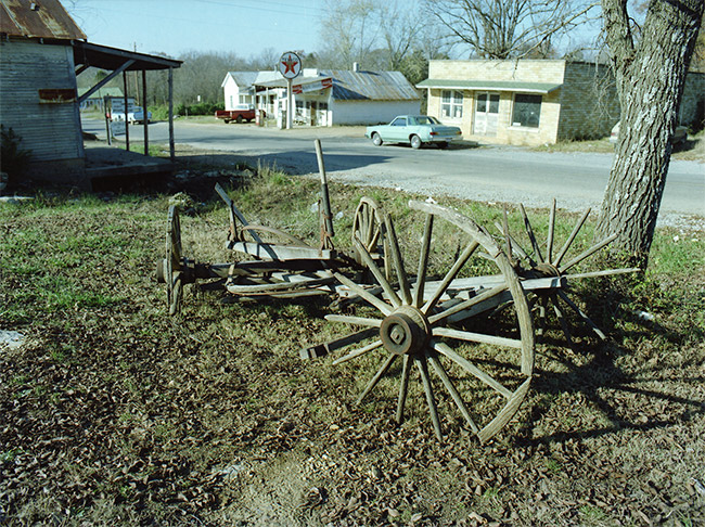 Remnants of wooden wagon and wagon wheel buried in the ground next to storefront on rural town road