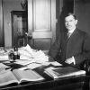 White man with mustache in suit and tie sitting at a table with papers and a telephone on it