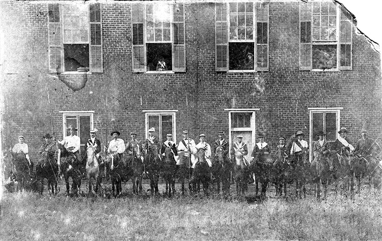 White men on horseback in front of multistory brick building