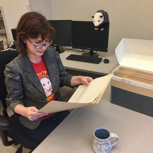 White woman with glasses sitting at desk with open folder in her hands with archival box and cup in front of her