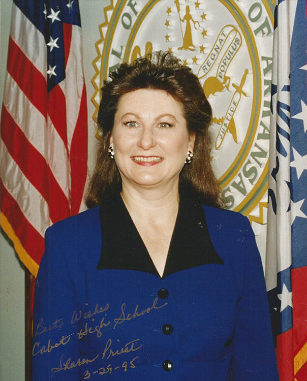 White woman with long hair smiling in blue top with flag and seal behind her