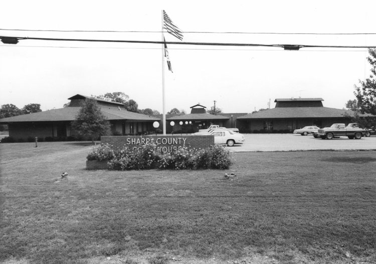 Single-story building with parking lot and flagpoles behind brick sign reading "Sharp County Courthouse"