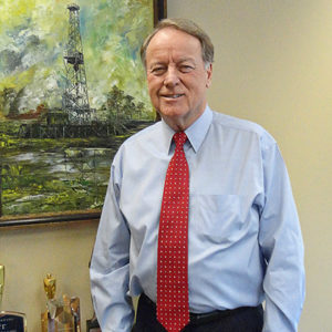 White man in shirt and tie smiling with painting of an oil derrick on the wall behind him above table with awards on it