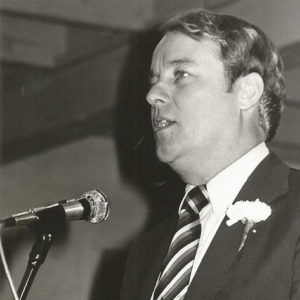 White man in suit and tie speaking at lectern