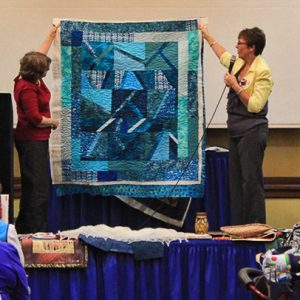 Two white women holding up a blue and white quilt while one speaks into a microphone