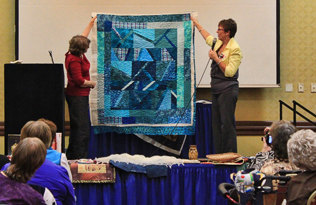 Two white women holding up a blue and white quilt while one speaks into a microphone