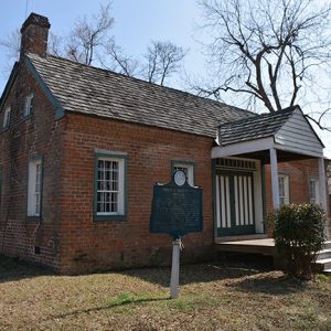 Single-story brick house with covered porch and historical marker sign in front yard