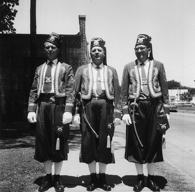 Three white men in Shriner's uniforms and hats with tassels with brick building behind them