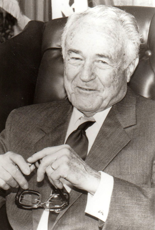 Old white man smiling in suit and tie seated in leather chair
