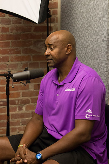 African American man in purple shirt and black shorts speaking into microphone