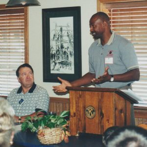 African-American man speaking at lectern with white man sitting beside him
