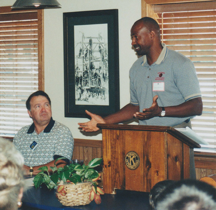 African-American man speaking at lectern with white man sitting beside him