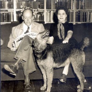 Middle-aged white man and woman siting on a couch with their dog and full bookshelves behind them