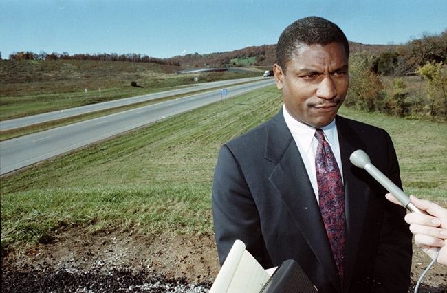 African-American man in suit and tie speaking into a microphone near interstate