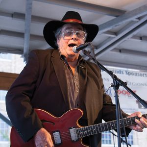 Older white man in hat and large glasses singing into a microphone while playing guitar