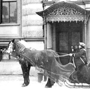 White woman and men in uniform on horse drawn sleigh outside  multistory building