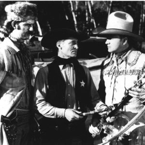 Three white men, one in raccoon hat, a sheriff, and a cowboy, examine hatchet bow and arrow