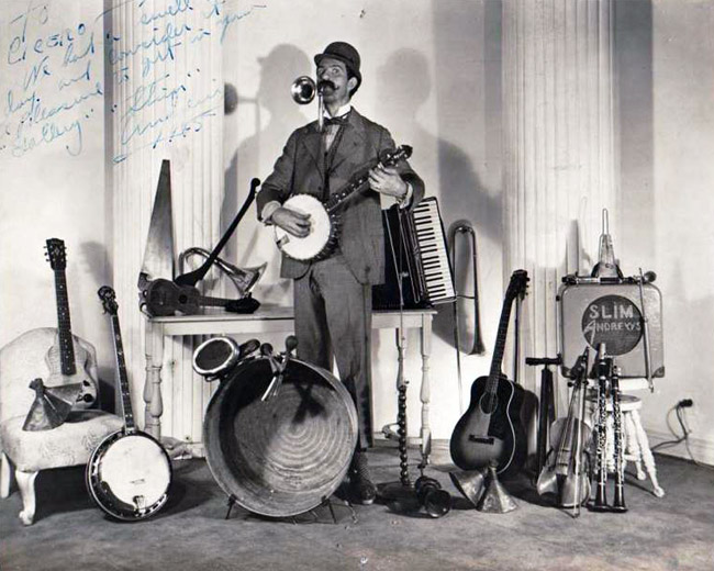 White man with mustache playing a horn and banjo surrounded by many musical instruments