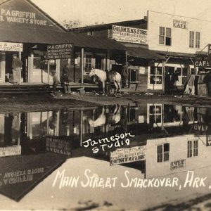 White men with horse drawn carriage and car outside single and multistory buildings reflected in flooded town street