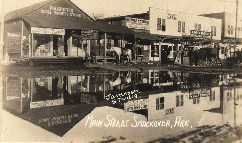 White men with horse drawn carriage and car outside single and multistory buildings reflected in flooded town street