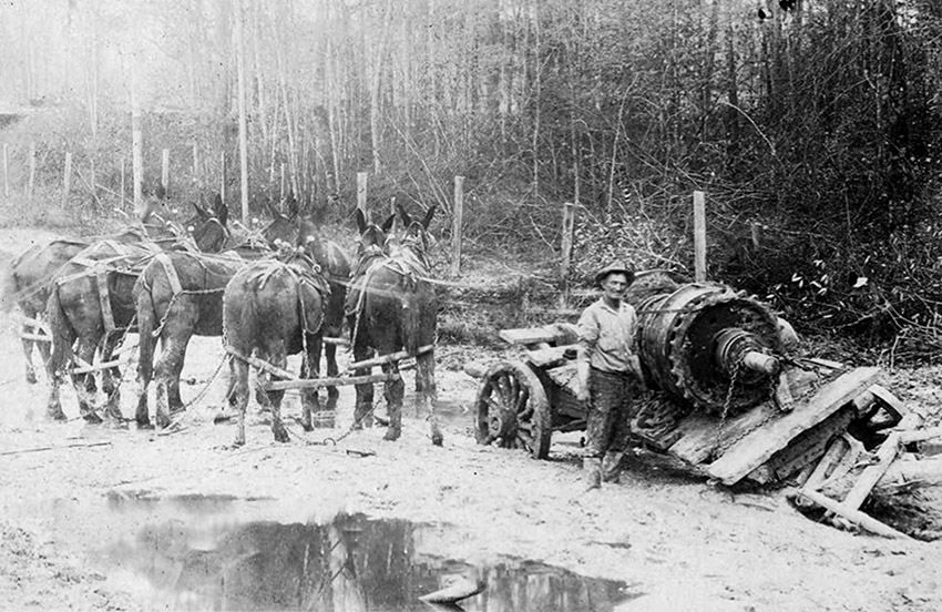 White man standing next to broken mule-drawn wagon with machinery on it on muddy road