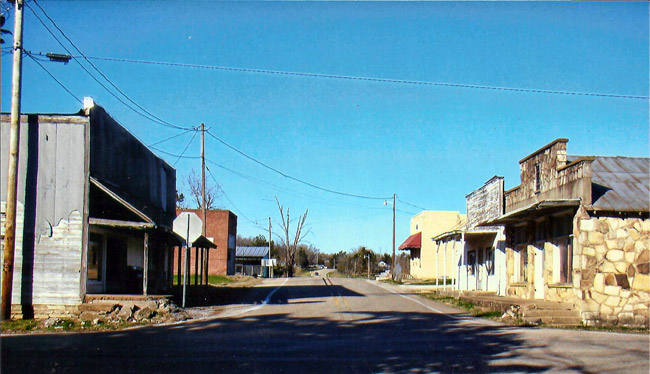 Street with storefront buildings on either side and power lines and blue sky