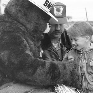 White man with mustache and cap with young white boy and person in Smokey the bear costume
