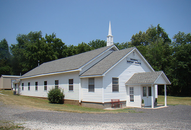 Single-story church building with steeple with gravel in front with sign saying "Snowball Baptist Church"