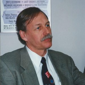 White man with mustache in suit and tie with poster behind him
