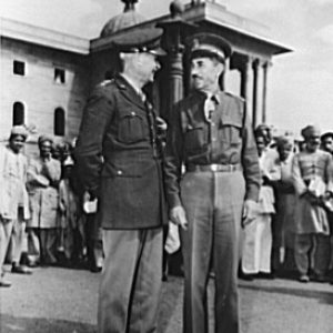 Two men in military uniforms talking with crowd and building behind them
