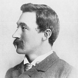 Profile view of white man with dark hair and mustache in suit jacket and tie