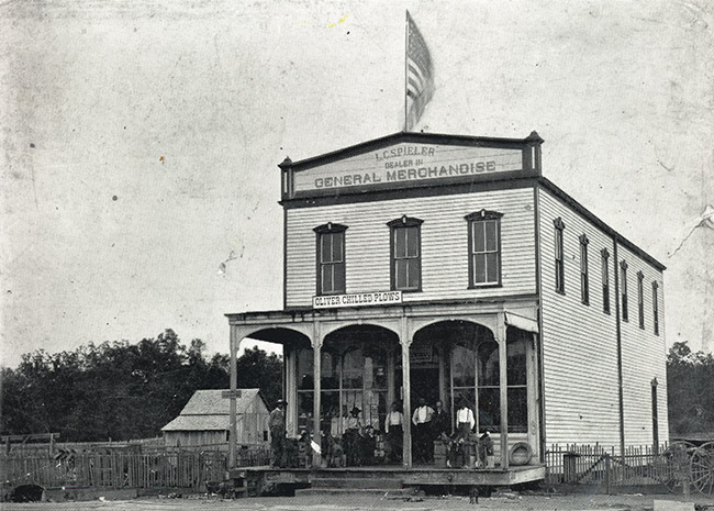 Group of white men outside two-story "General Merchandise" storefront with flag above it