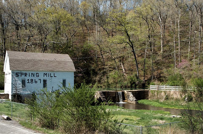 Barn with "Spring Mill 1867" written on its side and stone wall on creek with water flowing over it