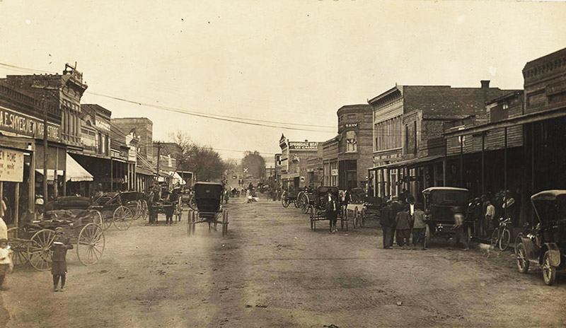 Cars and horse drawn carriages on street with multistory buildings on both sides