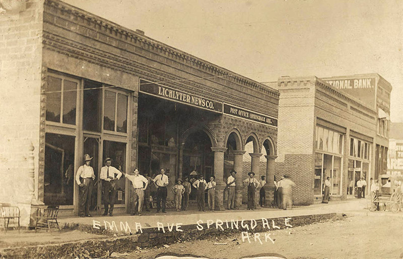 Group of white men and boys standing on sidewalk outside "Lichlyter News Company" building on town street