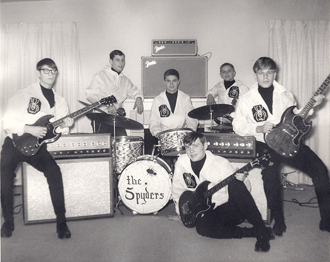 Six young white men in matching white jackets with guitars drums and amplifiers