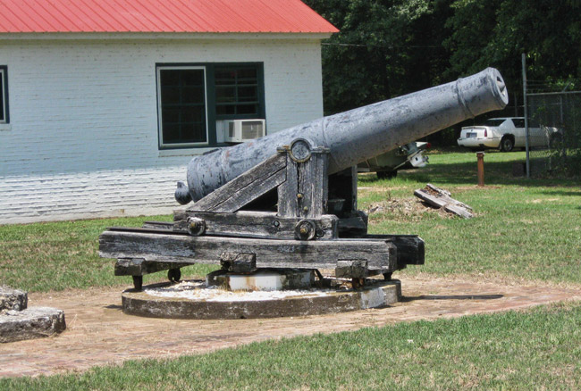 Cannon on round pedestal on concrete platform with single-story white building behind it
