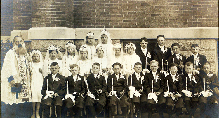Group of white children and in suits and dresses with white man in clerical robes outside brick building
