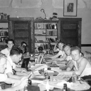 White men in casual dress studying at table with bookshelves on the wall and beds