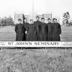 Group of white men in black religious garb with "St. John's Seminary" sign and large building in the far background