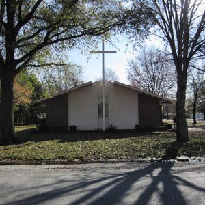 Single-story church building with large cross and trees on grass