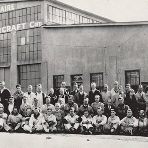 Large group of people sitting and standing in front of airplane hangar "Command-Aire"