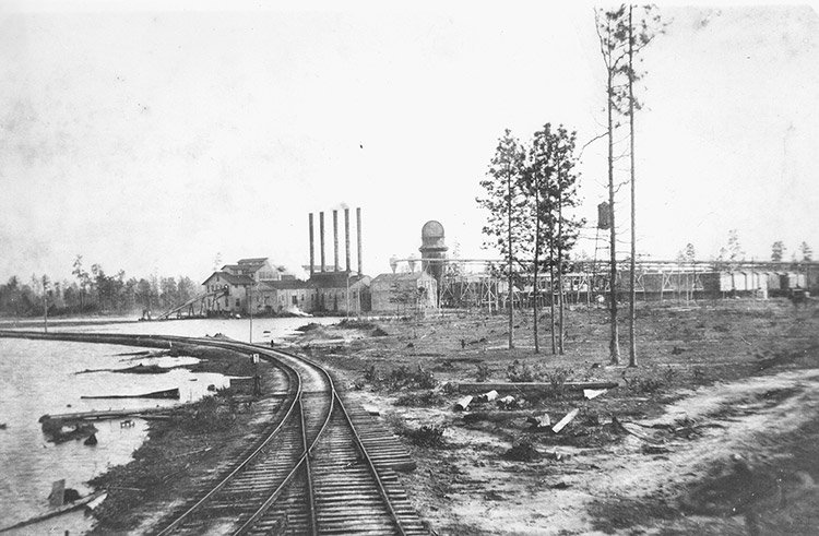 Multistory industrial buildings with four smokestacks and railroad tracks in the foreground