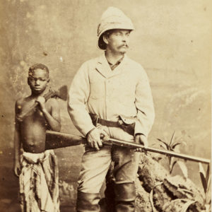 White man in pith helmet  holding rifle with shirtless black child behind him with rifle propped on his shoulder