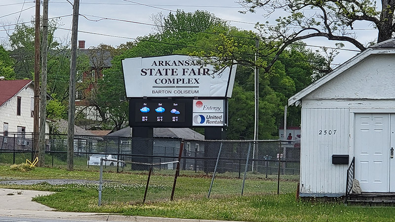 Multistory houses behind electronic Arkansas State Fair Complex sign with fence with single-story outbuilding in the foreground