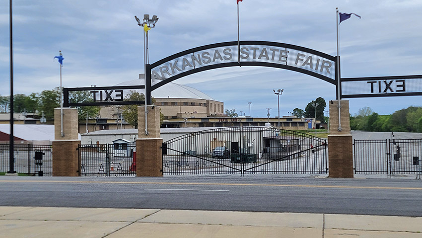 "Arkansas State Fair" arch sign over double gate with fence with single-story building and enclosed stadium in the background