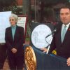 Older white woman standing with her designs on poster boards while a large white man speaks at a lectern