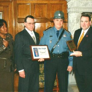 African-American woman standing with two white men and suits handing awards to white policeman in uniform