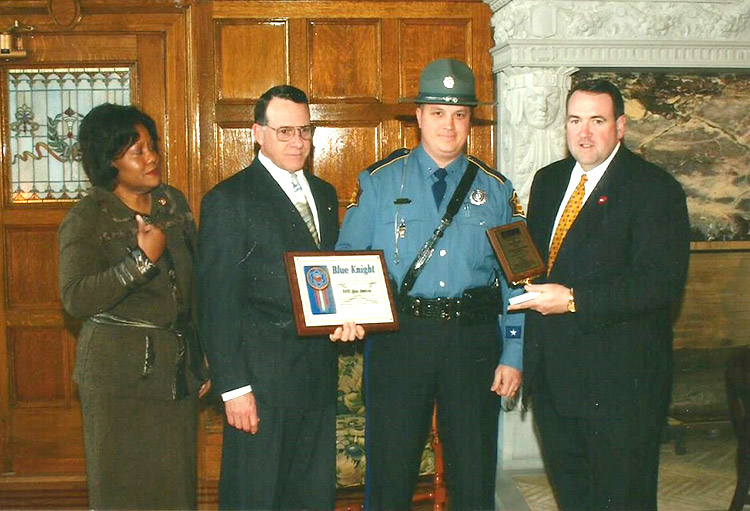 African-American woman standing with two white men and suits handing awards to white policeman in uniform